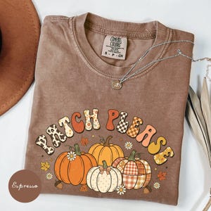 Peut inclure: Un t-shirt marron à manches courtes avec un col rond et une étiquette Comfort Colors. Le motif affiche le texte "PATCH PLEASE" dans des motifs variés au-dessus de trois citrouilles aux textures différentes, entourées de petites feuilles et fleurs.