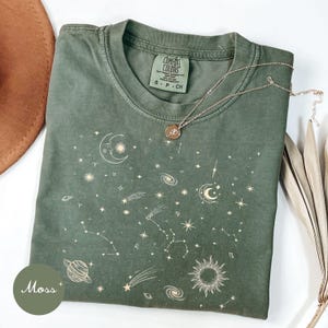Könnte beinhalten: Ein moosgrünes T-Shirt mit Rundhalsausschnitt und einem weißen himmlischen Grafikdruck aus Monden, Sonnen, Planeten, Sternen und Sternbildern. Das Innenetikett zeigt Comfort Colors und eine dünne goldfarbene Kette liegt auf dem Stoff.