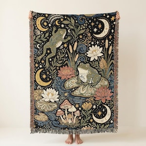 May include: A woven throw blanket featuring a black background with a pattern of green frogs, white and pink lotus flowers, mushrooms, and golden crescent moons. The design includes swirling water elements, cattails, and stars with fringed edges.