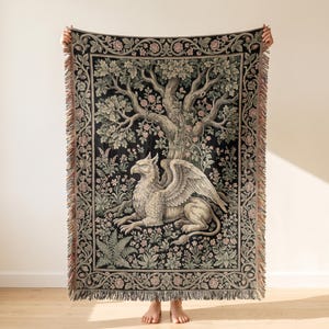 May include: A rectangular woven throw blanket featuring a central griffin sitting beneath a large tree. The design includes a black background with detailed floral motifs, green foliage, and a decorative border of roses, finished with fringed edges.
