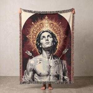 May include: A woven throw blanket featuring a depiction of a figure with wavy hair looking upward, surrounded by a golden circular halo against a deep red arched background. Three arrows are embedded in the chest area, and the blanket has fringe.
