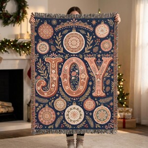 May include: A woven throw blanket featuring a dark blue background with the word JOY in large, ornate, floral-patterned letters. The design includes various circular Christmas ornaments, pine branches, and stars with fringed edges on all sides.