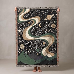 May include: A woven throw blanket featuring a black background with a celestial theme, including white constellations, planets, and a crescent moon. A large, flowing, multi-coloured ribbon in shades of green and tan curves across the centre above dark green mountain silhouettes at the bottom, finished with a fringed edge.