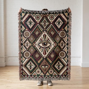 May include: A woven throw blanket featuring a repeating geometric pattern of diamonds and circles in shades of dark green, burgundy, and cream. The design includes multiple stylised eye motifs centred within the shapes, finished with a fringed edge.