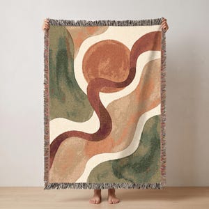 May include: A woven throw blanket featuring an abstract design with a large terracotta circle, a dark maroon winding line, and organic shapes in shades of sage green, cream, and tan. The blanket has a fringed edge on the top and bottom sides.