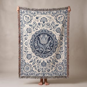 May include: A woven throw blanket featuring a cream-colored base with intricate dark blue floral and botanical patterns. The design includes a central circular medallion containing a large tulip, surrounded by scrolling vines, leaves, and pomegranates.