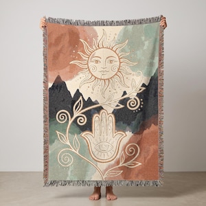 May include: A woven throw blanket featuring a sun with a face, a hamsa hand symbol, and a vine design. The background displays abstract shapes in shades of terracotta, sage green, and dark grey, with a fringed edge along the top and bottom.