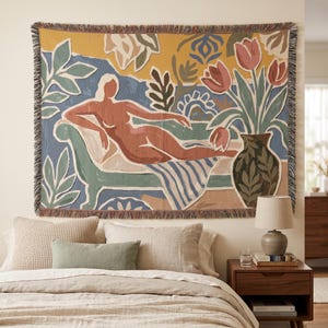 Puede incluir: Un tapiz de pared tejido que presenta una figura reclinada en tonos terracota sobre una chaise longue, rodeada de follaje verde estilizado y un jarrón de tulipanes rosas sobre un fondo amarillo mostaza, con un borde de flecos.