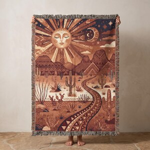 Pode incluir: Uma manta tecida retangular apresentando uma paisagem desértica com cactos, um caminho sinuoso e montanhas. A parte superior exibe um grande sol com um rosto humano e uma lua crescente contra um céu escuro. As bordas possuem acabamento em franjas.