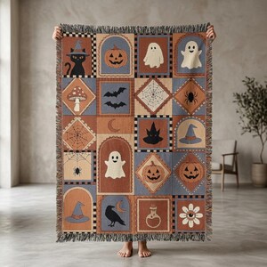 May include: A woven throw blanket featuring a grid of Halloween-themed illustrations including black cats, pumpkins, ghosts, bats, spiderwebs, spiders, mushrooms, witch hats, ravens, potion bottles, and flowers in shades of orange, blue, and cream.