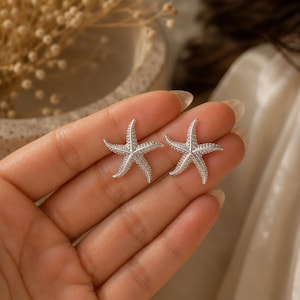 May include: A pair of silver-toned starfish earrings featuring a textured, dotted surface on each of the five arms. The starfish are shaped with a central point and radiating limbs, displaying a metallic finish that reflects light across the surface.