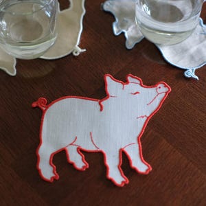 Puede incluir: Tres posavasos de tela con forma de cerdo, cada uno con un borde bordado distintivo. Un posavasos es blanco con borde rojo, otro es beige con borde blanco y el tercero es blanco con borde azul claro, cada uno sosteniendo un vaso de cristal.