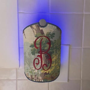 May include: A plug-in night light featuring a curved fabric cover with a printed landscape scene of trees and foliage. A large, embroidered red letter B is centered on the front. The device emits a bright blue glow from behind the cover.