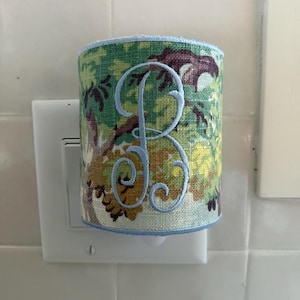 May include: A cylindrical plug-in night light cover featuring a green, yellow, and brown forest-themed fabric pattern. The cover is accented with a light blue piped trim and displays a large, embroidered light blue script letter B centered.
