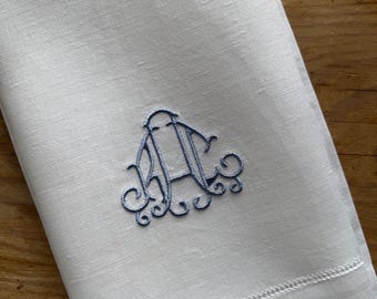 Pode incluir: Um guardanapo de linho branco com um monograma decorativo bordado em azul no centro. O tecido tem uma textura visível e um detalhe de bainha aberta ao longo da borda inferior, exibindo um design limpo e simples.