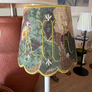 Pode incluir: Um pequeno abajur c&oacute;nico com uma estampa floral texturizada e multicolorida em tons de verde, castanho e roxo. O abajur &eacute; acentuado com um grande monograma amarelo bordado com as letras M e R, finalizado com uma borda festonada.