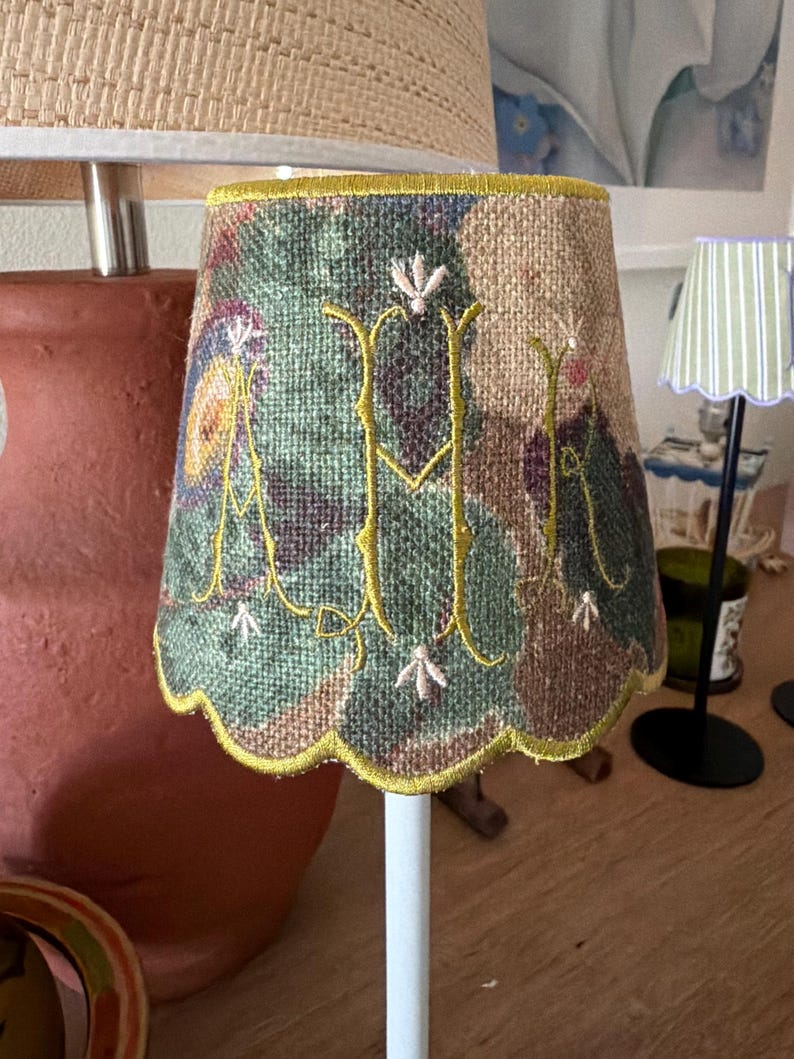 Pode incluir: Um pequeno abajur c&oacute;nico com uma estampa floral texturizada e multicolorida em tons de verde, castanho e roxo. O abajur &eacute; acentuado com um grande monograma amarelo bordado com as letras M e R, finalizado com uma borda festonada.