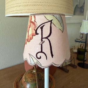 Pode incluir: Um pequeno abajur de tecido com borda festonada em tom rosa suave, apresentando uma letra R grande bordada em marrom escuro com uma fonte cursiva decorativa. O abajur &eacute; adornado com um padr&atilde;o floral em tons suaves de laranja, verde e creme.