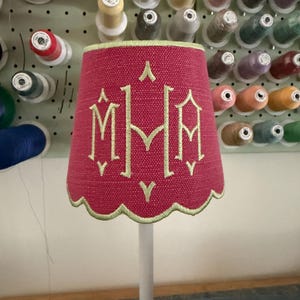 Pode incluir: Um abajur cil&iacute;ndrico com um exterior de tecido rosa choque texturizado e uma borda inferior recortada. O abajur apresenta um monograma bordado em verde claro com as letras M, M e A, acentuado por pequenos detalhes de costura verde.