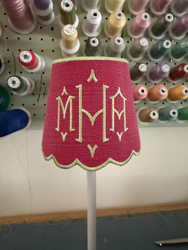 Pode incluir: Um abajur cil&iacute;ndrico com um exterior de tecido rosa choque texturizado e uma borda inferior recortada. O abajur apresenta um monograma bordado em verde claro com as letras M, M e A, acentuado por pequenos detalhes de costura verde.