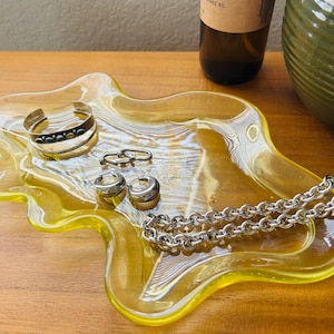 May include: A translucent yellow glass trinket tray with an irregular, wavy edge. The tray holds a silver-toned cuff bracelet with small dark stones, two small silver hoop earrings, and a thick silver-toned chain link necklace.