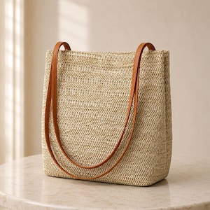Op de afbeelding: Een rechthoekige tote bag gemaakt van geweven beige stro met een opvallend visgraatpatroon. De tas heeft twee lange, dunne, gladde, lichtbruine schouderbanden die zijn bevestigd aan de bovenrand van de gestructureerde, open tas.
