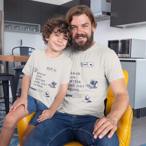 Op de afbeelding: Een beige t-shirt met korte mouwen en een donkerblauwe grafische print van een computer, een muis en een laadbalk met de tekst "ERROR 404: BETTER DAD NOT FOUND." Het ontwerp bevat ook een klein kubus- en wereldbolpictogram.