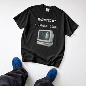 以下が含まれることがあります： 黒の半袖Tシャツ。白い文字で「HAUNTED BY >LEGACY CODE_」とプリントされ、その下に幽霊が映るベージュのヴィンテージ風コンピュータモニターのグラフィックが描かれています。クルーネックで標準的なフィット感です。
