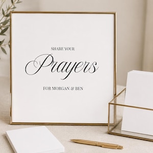 May include: A square sign in a thin gold frame with white background and black text that reads SHARE YOUR Prayers FOR MORGAN & BEN. Next to the sign is a glass box containing a stack of white cards, a gold pen, and a small white notepad.