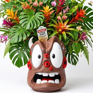 Pode incluir: Um vaso em forma de monstro castanho com vários olhos, boca aberta e dentes brancos, cheio de plantas tropicais vibrantes, incluindo bromélias cor de laranja e folhas de monstera. Uma etiqueta diz WARNING: EXTREME GROWTH.