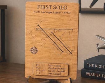 Puede incluir: Una placa rectangular de madera con un mapa grabado del aeropuerto de North Las Vegas, el texto FIRST SOLO y una rosa de los vientos. Una placa de madera más pequeña muestra detalles del vuelo, incluyendo la fecha 30 de octubre de 2022 y el avión C172N.