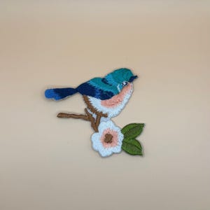 May include: An embroidered patch featuring a small bird with teal, dark blue, and light pink plumage perched on a brown branch. Below the branch is a white flower with a pink center and two green leaves, all rendered in detailed thread work.
