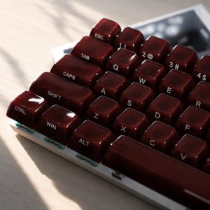 Könnte beinhalten: Eine Computertastatur mit glänzenden, tiefroten Tastenkappen und weißer Beschriftung. Die Tasten tragen Standardbezeichnungen wie ESC, TAB, CAPS, SHIFT, CTRL, WIN, ALT sowie alphanumerische Zeichen. Die Tastatur hat einen weißen Rahmen.