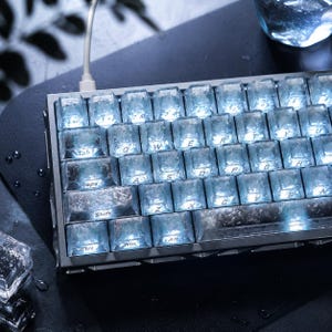 Könnte beinhalten: Eine mechanische Tastatur mit durchscheinenden, eisartigen Tastenkappen in einer texturierten, gefrosteten Optik. Die Tasten sind mit hellem, weißem Licht hinterleuchtet und zeigen das Standard-QWERTY-Layout. Ein weißes Kabel ist oben angeschlossen.