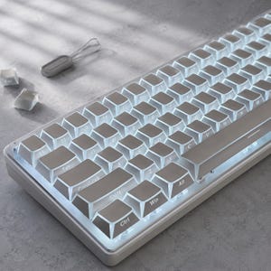 Könnte beinhalten: Eine weiße mechanische Tastatur mit hintergrundbeleuchteten Tasten und kompaktem Layout. Die Tasten zeigen die Standard-QWERTY-Beschriftung. Ein kleiner grauer Tastenkappen-Abzieher und zwei lose weiße Tastenkappen liegen oben links.