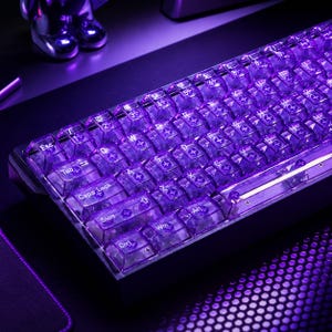 Könnte beinhalten: Eine mechanische Tastatur mit durchscheinenden violetten Tastenkappen und weißer Beschriftung. Die Tasten werden durch eine leuchtend violette Hintergrundbeleuchtung erhellt, die die klare Kunststoffkonstruktion und das QWERTY-Layout betont.