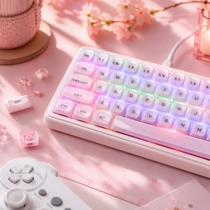 Könnte beinhalten: Eine kompakte mechanische Tastatur mit weißem Rahmen und durchscheinenden weißen Tastenkappen mit schwarzer Beschriftung. Die Tastatur verfügt über eine integrierte RGB-Hintergrundbeleuchtung in Pastellfarben. Ein weißer Gamecontroller ist im Vordergrund zu sehen.