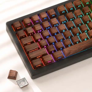 May include: A mechanical keyboard featuring dark brown wood grain keycaps with white lettering and integrated RGB backlighting. The keyboard includes a black frame, and two additional wood grain keycaps are positioned separately nearby.