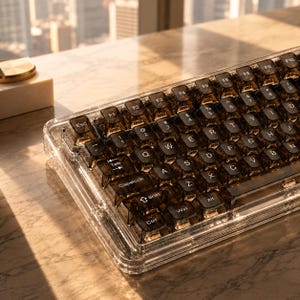 May include: A mechanical keyboard featuring a transparent clear acrylic frame and dark translucent keycaps with white lettering. The QWERTY layout includes standard function keys, a tab key, a caps lock key, and a shift key, all visible in bright light.