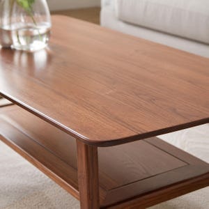 May include: A rectangular coffee table crafted from medium-toned brown wood with a smooth, polished surface. The table features rounded corners and a lower shelf for storage, supported by four sturdy legs, showcasing a visible natural wood grain.