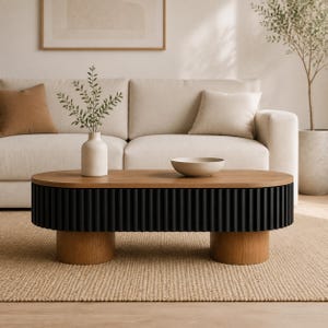 May include: An oval coffee table featuring a light wood top and a black fluted base supported by two cylindrical light wood legs. A light-colored ceramic vase with green foliage and a shallow beige bowl are placed on the table surface.