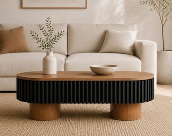 May include: An oval coffee table featuring a light wood top and a black fluted base supported by two cylindrical light wood legs. A light-colored ceramic vase with green foliage and a shallow beige bowl are placed on the table surface.