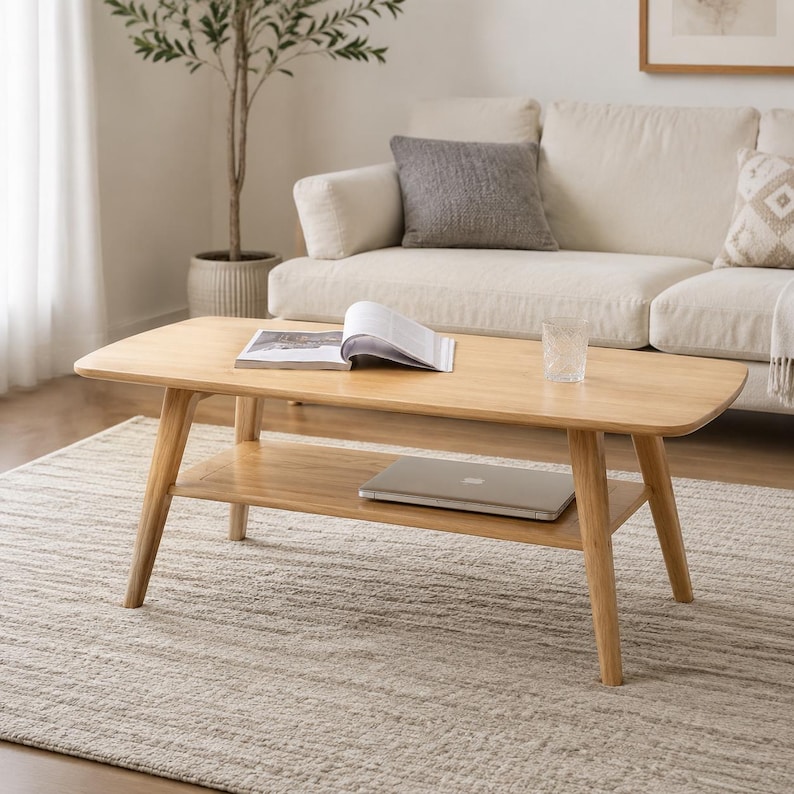May include: A light-toned wooden coffee table featuring a rectangular top with rounded corners and four angled legs. The table includes a lower shelf for storage, currently holding a closed silver laptop, while an open book rests on the top surface.