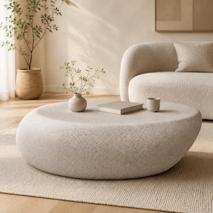 May include: A low, round, pebble-shaped coffee table with a textured, light beige stone-like finish. The surface holds a small, smooth, light-colored ceramic vase containing delicate green sprigs, a closed book, and a small matching cup.