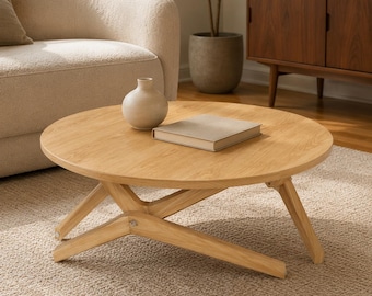 May include: A round light-toned wooden coffee table with a smooth surface and a unique intersecting leg design. A small, matte beige ceramic vase and a closed, light-colored rectangular book are placed on top of the table's flat surface.