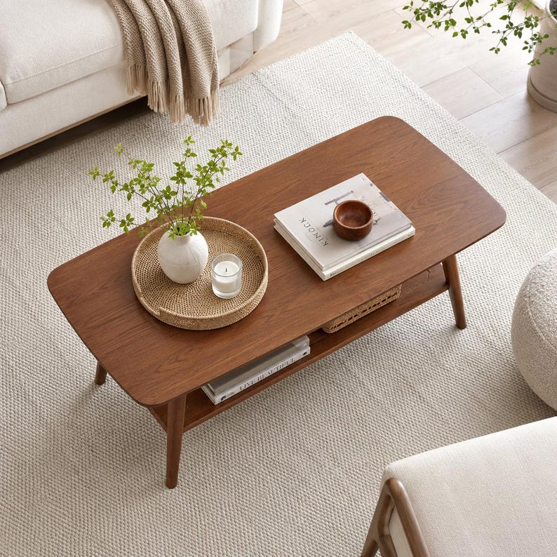 May include: A rectangular wooden coffee table with rounded corners and a lower shelf. The tabletop holds a round woven tray with a white vase containing green stems and a glass candle, plus a stack of books topped with a small wooden bowl.