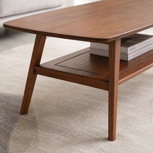 May include: A rectangular coffee table crafted from dark brown wood with a smooth, polished surface. The table features four tapered legs and a lower shelf that holds two stacked books with the visible text Nordic and Kinfolk on the spines.