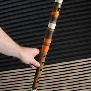 May include: A cylindrical bamboo flute featuring a series of finger holes along its length. The instrument is decorated with alternating bands of white, orange, and dark brown thread wrapped around the body at various intervals along the shaft.