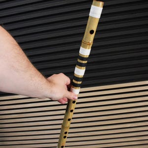 Op de afbeelding: Een bamboe fluit met een natuurlijke lichtbruine afwerking en meerdere vingergaten. Het instrument is versierd met afwisselende banden van wit en zwart garen die op verschillende intervallen strak om het cilindrische lichaam zijn gewikkeld.