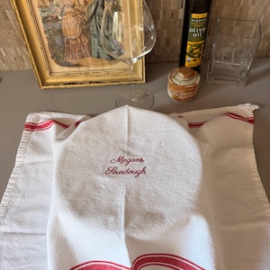 May include: A white kitchen towel with red stripes along the edges, featuring the embroidered red text "Megan's Sourdough" in a cursive script. The towel is draped over a rounded object, creating a soft, folded appearance on the surface.